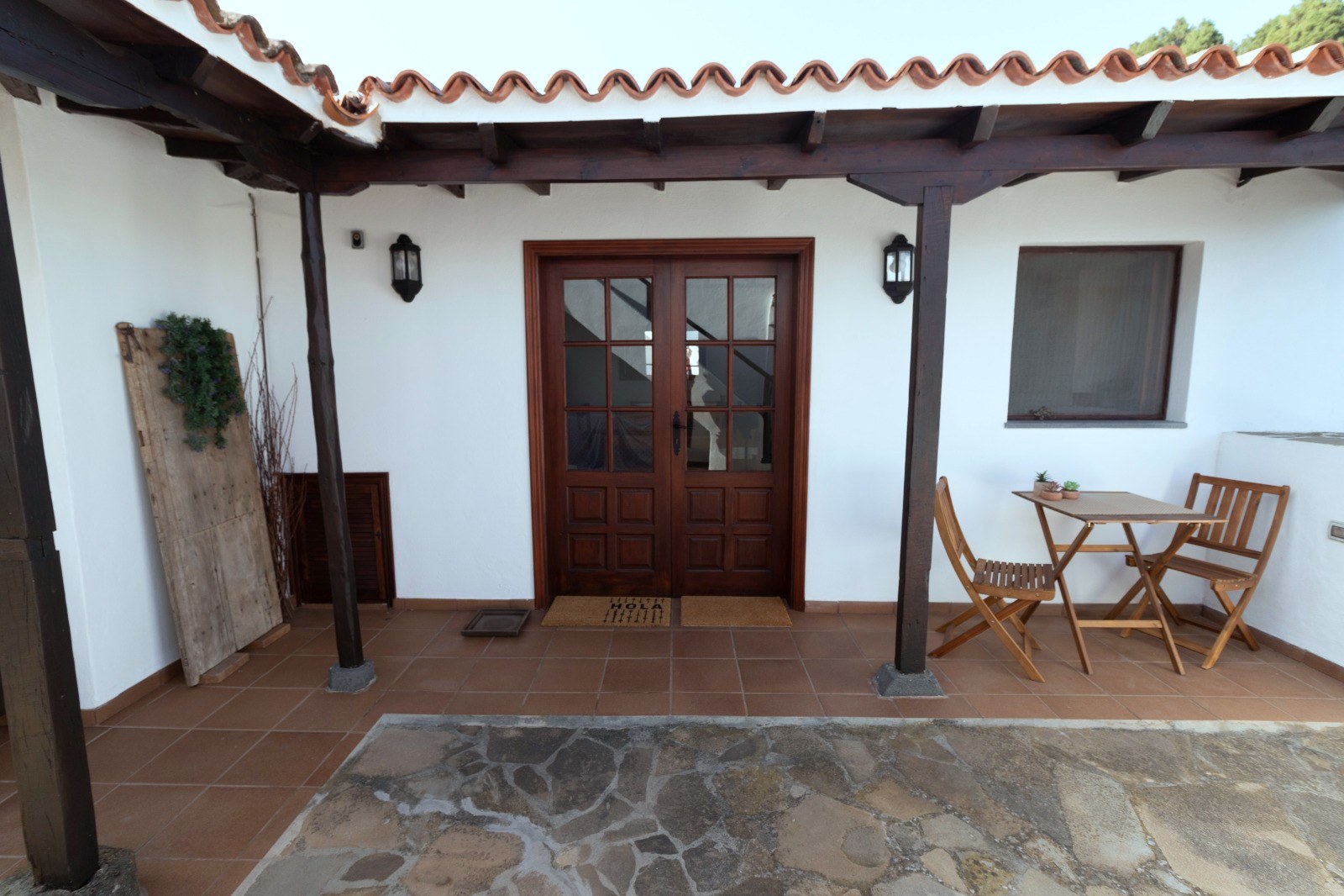 Entrada principal de Finca Julia bajo la pérgola de madera, con puerta acristalada, felpudo y una mesa para dos en el patio de losa